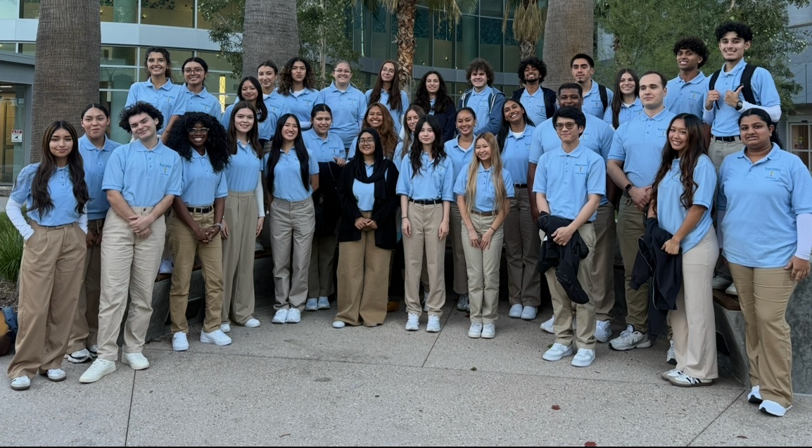 Large COPE Health Scholars cohort in blue polos gathered outside a modern healthcare and academic setting